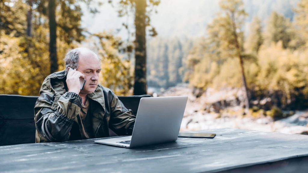 Person mit Outdoor-Jacke sitzt an einem Holztisch im Freien vor einem geöffneten Laptop, umgeben von Wald und Bergen.
