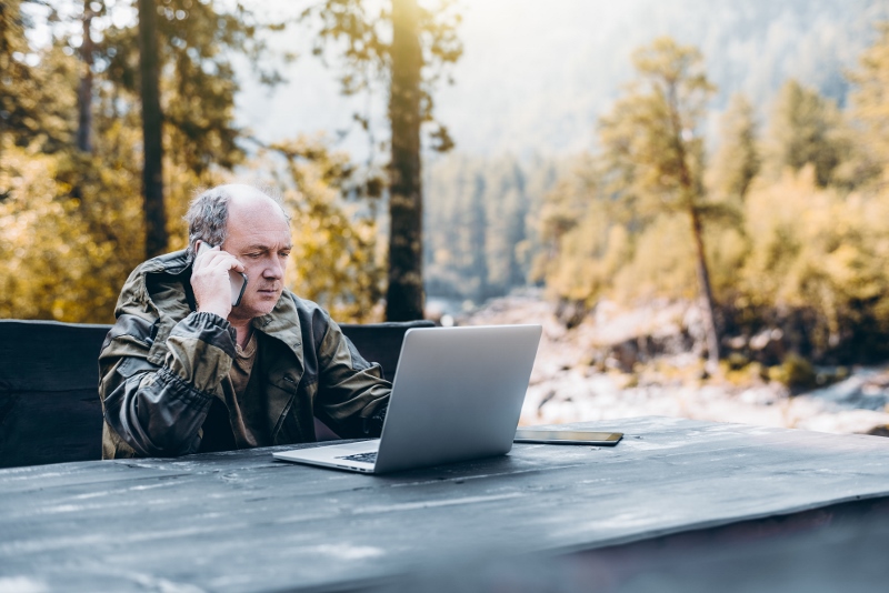 Personne en veste d'extérieur assise à une table en bois en plein air devant un ordinateur portable ouvert, entourée de forêt et de montagnes.